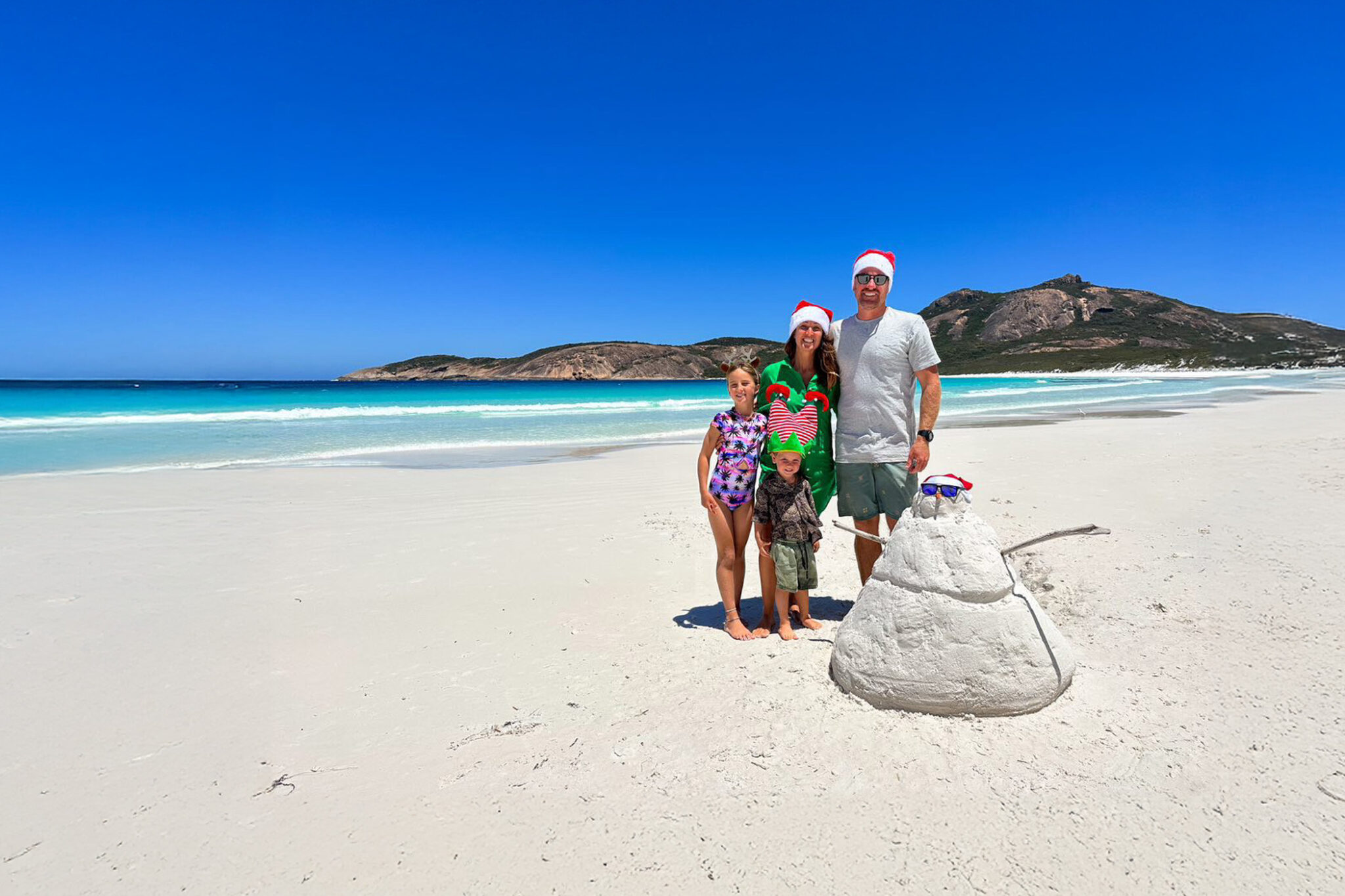 A family on a beach wearing Christmas items standing next to a sandman that looks like a snowman