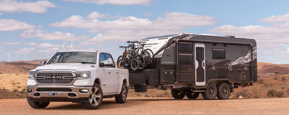 A white RAM pickup towing a black Wonderland RV off-road caravan with mountain bikes mounted on the front, set against a remote, arid landscape with wind turbines in the background.