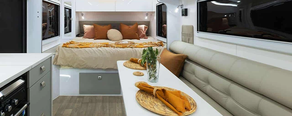 Modern caravan interior featuring a queen bed with warm-toned bedding, a soft leather lounge, a white dining table set with woven placemats, and a compact kitchen.