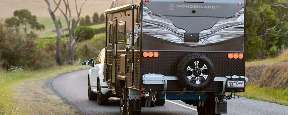 A Wonderland RV off-road caravan being towed by a white SUV along a winding country road at sunset, with rolling hills and trees in the background.
